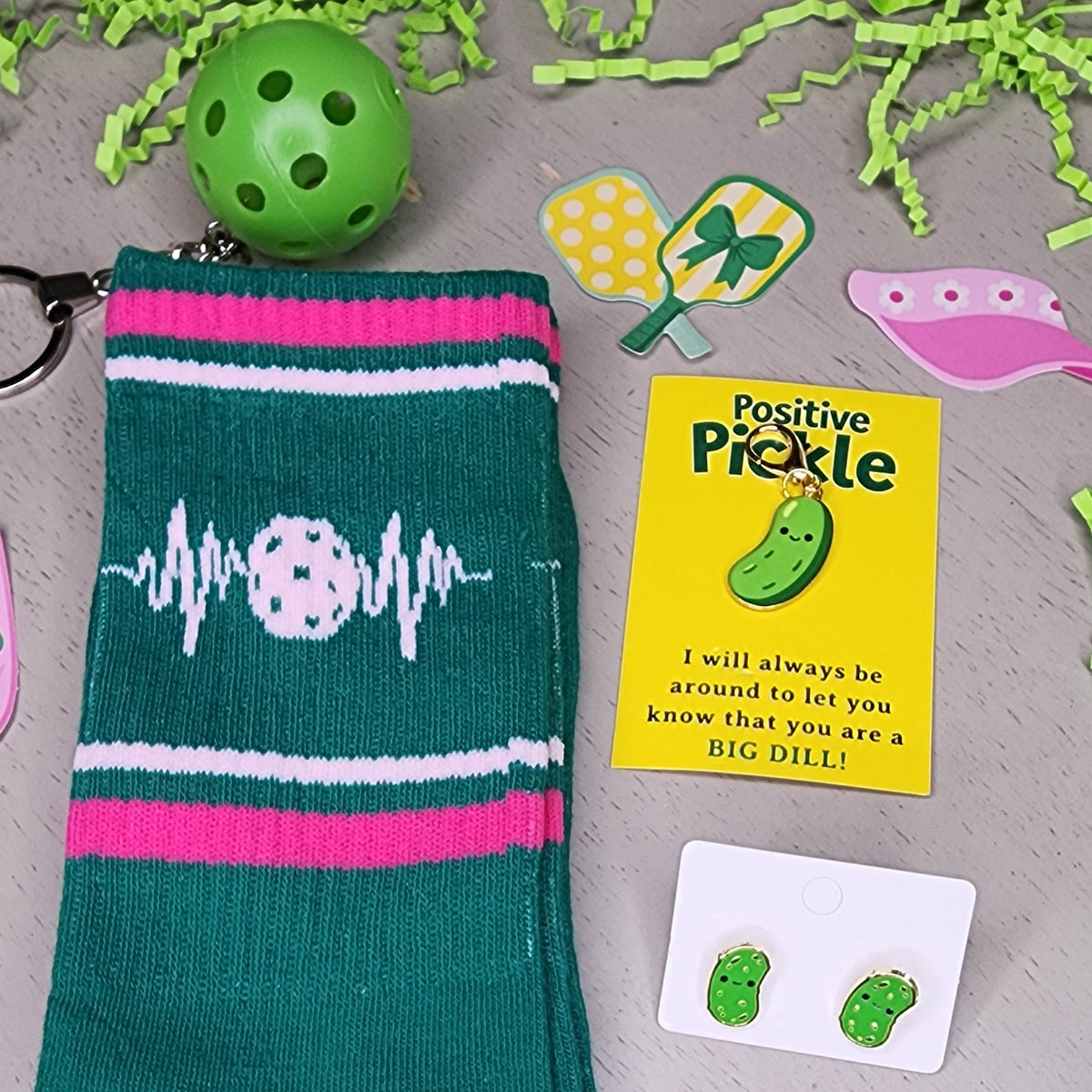 A flat-lay of a novelty pickleball-themed sock set on a gray surface with green paper shred. The set includes a dark teal sock with pink and white stripes, a pickleball graphic, and a white toe, alongside a green pickleball, a yellow 'Positive Pickle' card, green stud earrings, and a 'Pickleball Club' sticker.