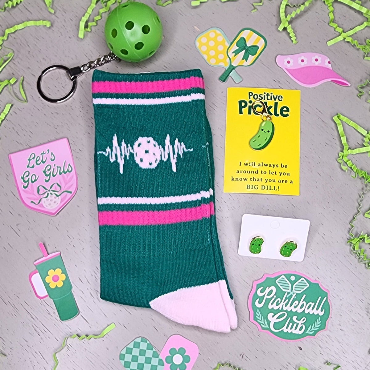 A flat-lay of a green and pink pickleball themed crew sock with a white heartbeat line and soccer ball graphic, accompanied by a green soccer ball keychain, a yellow 'Pickle' sticker, and other matching green and pink stickers on a gray surface.